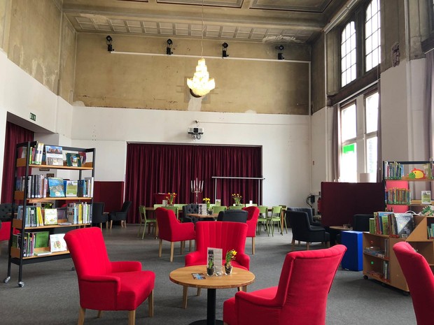 Neu gestalteter Innenraum des Bahnhofs Löhne mit roten Sesseln, kleinen runden Holztischen, Deckenleuchte und Bücherregalen.