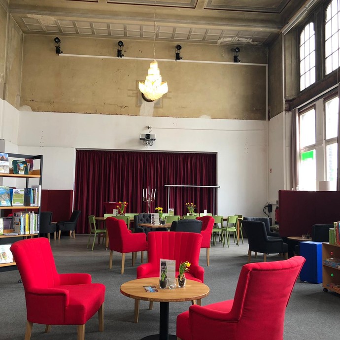 Neu gestalteter Innenraum des Bahnhofs Löhne mit roten Sesseln, kleinen runden Holztischen, Deckenleuchte und Bücherregalen. (vergrößerte Bildansicht wird geöffnet)