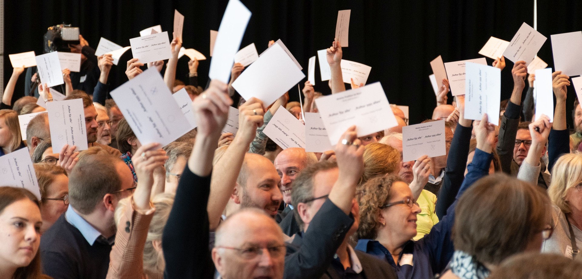 Die Teilnehmer:innen der Westfälischen Kulturkonferenz heben die Hände mit einem Stimmzettel für eine Abstimmung.