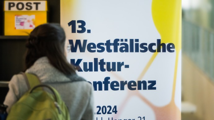 Eine Person steht mit dem Rücken zur Kamera vor dem Plakat der Kulturkonferenz.